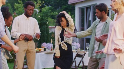 Beautiful brunette woman dancing joyfully with hand in hair surrounded by friends. People moving and smiling. Standing near dinner table. Celebrating lively evening outdoors with laughter and music. - Powered by Adobe