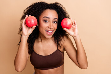 Cheerful young woman holding two bright apples against a neutral background embodying fun, health,...