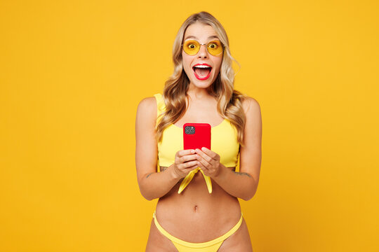 Young shocked surprised fun beautiful sexy woman wear bikini swimsuit rest near hotel pool hold in hand use mobile cell phone isolated on plain yellow background. Summer vacation sea sun tan concept.