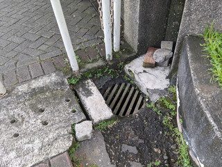 A damaged storm drain with broken concrete surrounds it, next to a sidewalk and metal pipes.