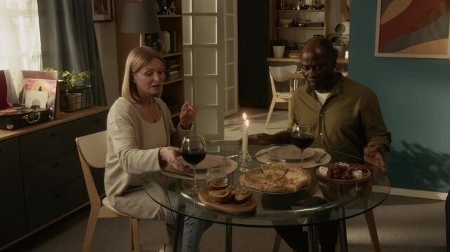 Multiethnic mature couple starting romantic dinner sitting at round table with candles, baked pears, pizza and appetizers, they clinking glasses filled with red wine