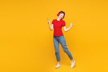 Fototapeta premium Full body young woman she wearing red t-shirt casual clothes listen to music in headphones holding in hand use mobile cell phone isolated on plain yellow background studio portrait. Lifestyle concept.