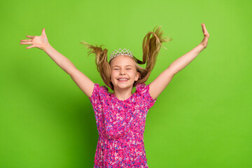 Smiling girl with raised hair wearing crown and dress posing joyfully against a green background