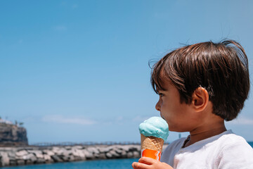 A young boy is holding a blue ice cream cone in his hand. The sky is clear and blue, and the ocean is in the background. The boy appears to be enjoying his ice cream as he looks out at the water