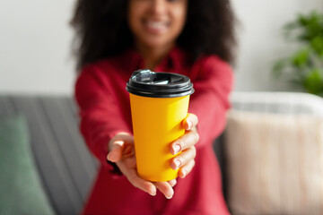 Smiling African American woman offering yellow disposable coffee cup, selective focus, mockup