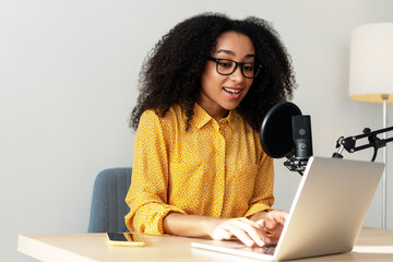 Smiling cheerful African American woman recording podcast from home using laptop and microphone