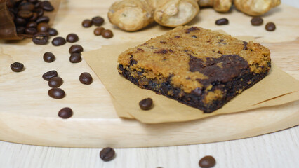 Blondie and brownie mix bar resting on baking paper on a wooden board, with coffee beans and ginger root scattered around, creating a tempting presentation of flavors