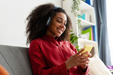 Beautiful African American woman listening to music on headphones and using smartphone at home