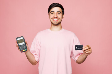 Young man he wearing t-shirt casual clothes hold wireless modern bank payment terminal to process acquire credit card isolated on plain pastel light pink background studio portrait. Lifestyle concept.