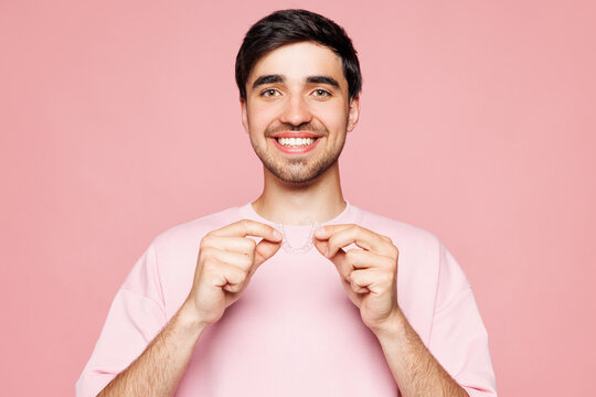 Fototapeta Young smiling happy man wears t-shirt casual clothes hold in hand invisible transparent aligners, silicone invisalign dental braces isolated on plain pastel light pink background. Lifestyle concept.