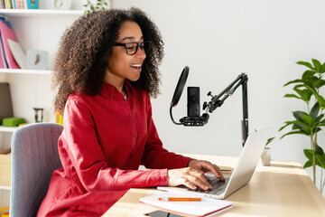 Young African American woman wearing eyeglasses recording podcast using laptop and microphone
