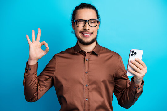 Young man in brown shirt holding smartphone and making OK hand gesture against turquoise background
