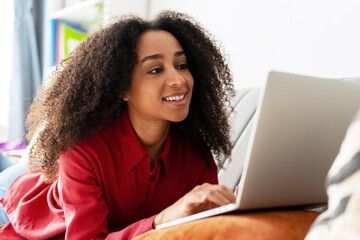Smiling African American woman lying on sofa using laptop, working remotely from home