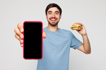 Young man wear blue t-shirt casual clothes eat show burger hold use blank screen area mobile cell...