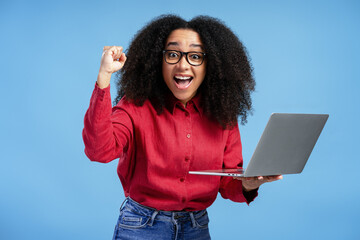 Excited young African American woman celebrating success with laptop on blue background