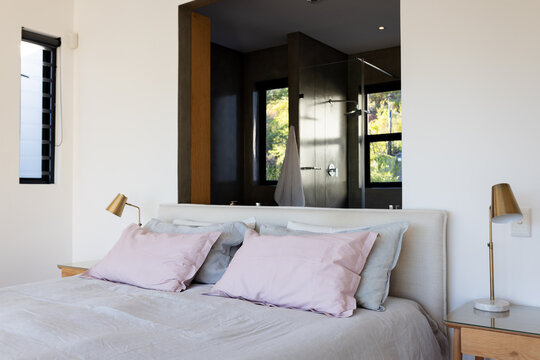 Queen bed resting with gray duvet, pillows in master bedroom opening into glass shower with towel