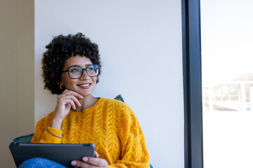 African American woman wearing glasses and mustard sweater sitting on window seat holding tablet