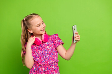 Smiling young girl with headphones and phone against green background, posing cheerfully in bright...