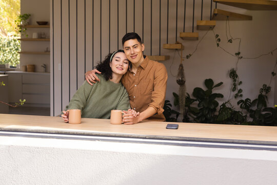 Diverse couple holding beige mugs at countertop in home kitchen with smartphone and wood staircase - Powered by Adobe
