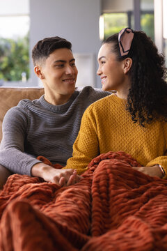 Couple sitting at home on sofa wearing headband and snuggling under rust-colored blanket by window