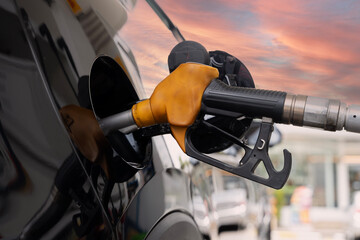 Fuel nozzle refueling black car at gas station with sunset sky reflection on surface, close up...