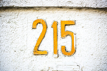 Bold yellow house number 215 painted on a rough white stucco wall. The textured background and...