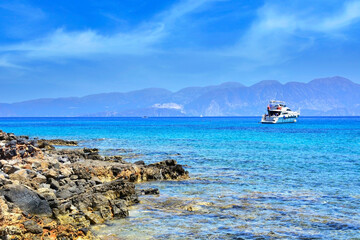 A boat at anchor near the rocky shore of the Mediterranean Sea with turquoise water, rocky hills in the background.
