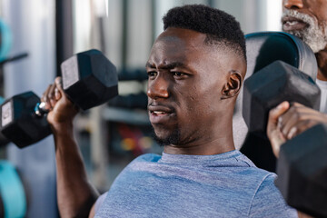 African American man pressing hex dumbbells on gym bench with senior trainer assisting