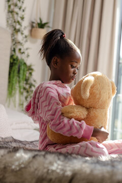 African American child wearing pink zigzag pajamas and holding teddy bear on bed in bedroom