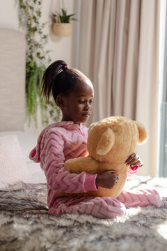 African American girl in pink hooded onesie hugging plush teddy bear on bed near potted plant