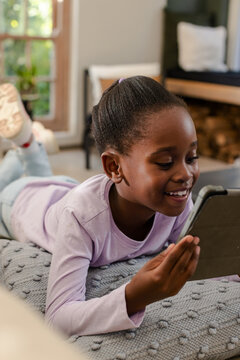 African American school-aged girl lying on gray blanket in living room holding tablet computer