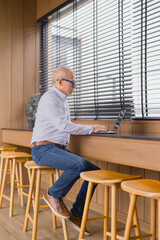 Focused mature businessman using laptop computer while sitting on stool at high table in...