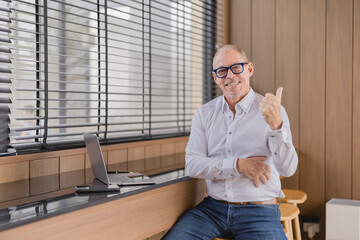 Smiling businessman gesturing thumbs up sitting on a stool in a modern office with laptop on the...