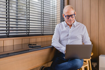 Smiling senior businessman working on laptop in modern office with window blinds, sitting on chair...