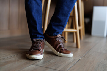 Close-up of a man's feet wearing stylish brown leather sneakers and blue jeans, sitting on a wooden...