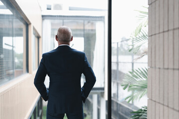 Senior executive gazing out the window of a modern office building, reflecting on the future of the...