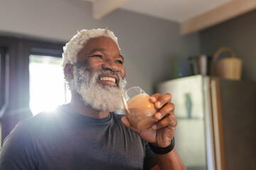 African American senior man smiling wearing smartwatch, holding frothy coffee mug in sunlit kitchen
