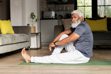 Senior African American man stretching on green yoga mat in living room checking fitness tracker