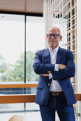 Mature businessman standing confidently in a modern office setting, wearing glasses and a suit. The...