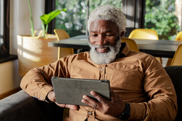 Senior African American man wearing smartwatch sitting on sofa using black tablet in living room