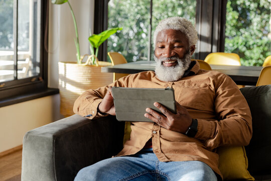Senior African American man sitting on sofa at home using tablet with green plant beside window