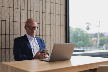 Senior manager working with mobile phone and computer sitting at desk in contemporary workplace, businessman using smartphone and laptop in modern office