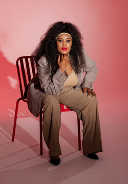 African American woman sitting in studio on red metal chair wearing blazer leaning touching chin