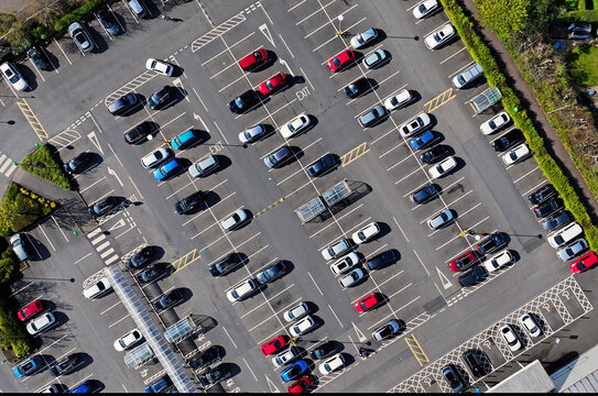 Aerial drone view of multiple coloured cars and vehicles parked in white painted spaces in a large town centre pay and display car park in UK
