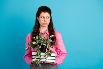 Young woman holding books and a plant against a blue background expressing thoughtful emotions in a...