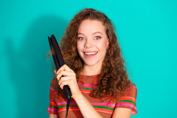 Fototapeta premium Smiling young woman with curly hair holding a straightener on a vibrant turquoise background in casual striped attire