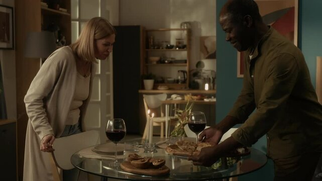 Black man placing plate with pizza on round glass table and looking at it with enjoyment, happy Caucasian woman blowing off candle and coming up to him