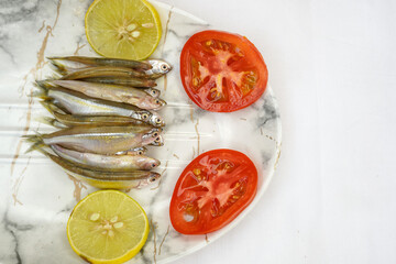 Fresh raw small fish on white plate with lemon and tomato on white textured wooden background, top view. Culinary seafood concept. copy space.
