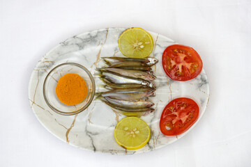 Fresh raw small fish decorated with lemon slices and tomato slices, glass bowl with turmeric powder on the white background. Healthy food concept, top view.