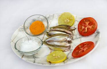Fresh raw small fish decorated with lemon slices and tomato slices, glass bowl salt and turmeric powder on the white background. Healthy food concept, top view.